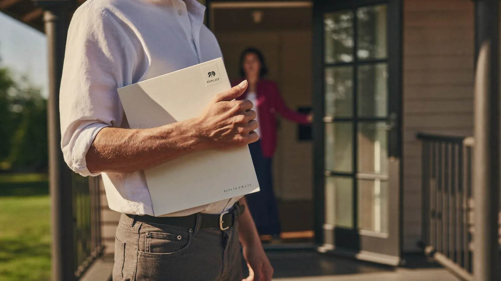 A salesperson on a front porch holding a folder, talking with a homeowner in the doorway in warm afternoon light.