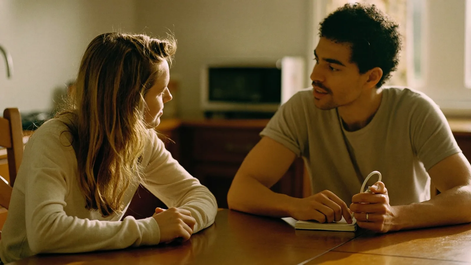 Two people in a warm conversation across a kitchen table, one taking notes in a notebook.