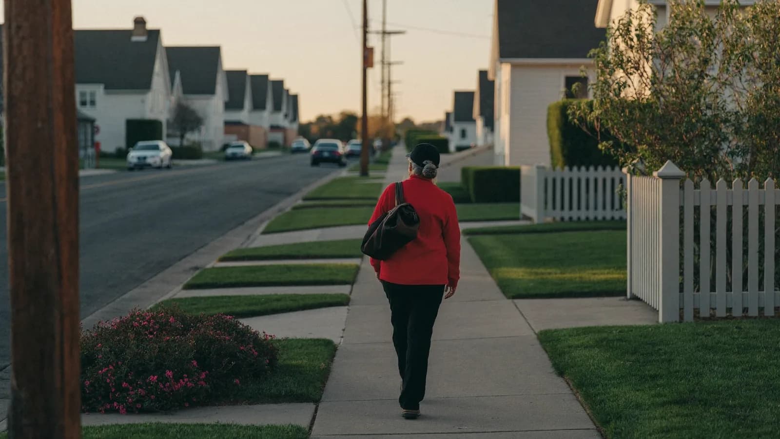 A quiet suburban street at golden hour with long shadows and a door-to-door salesperson walking the sidewalk with a bag.