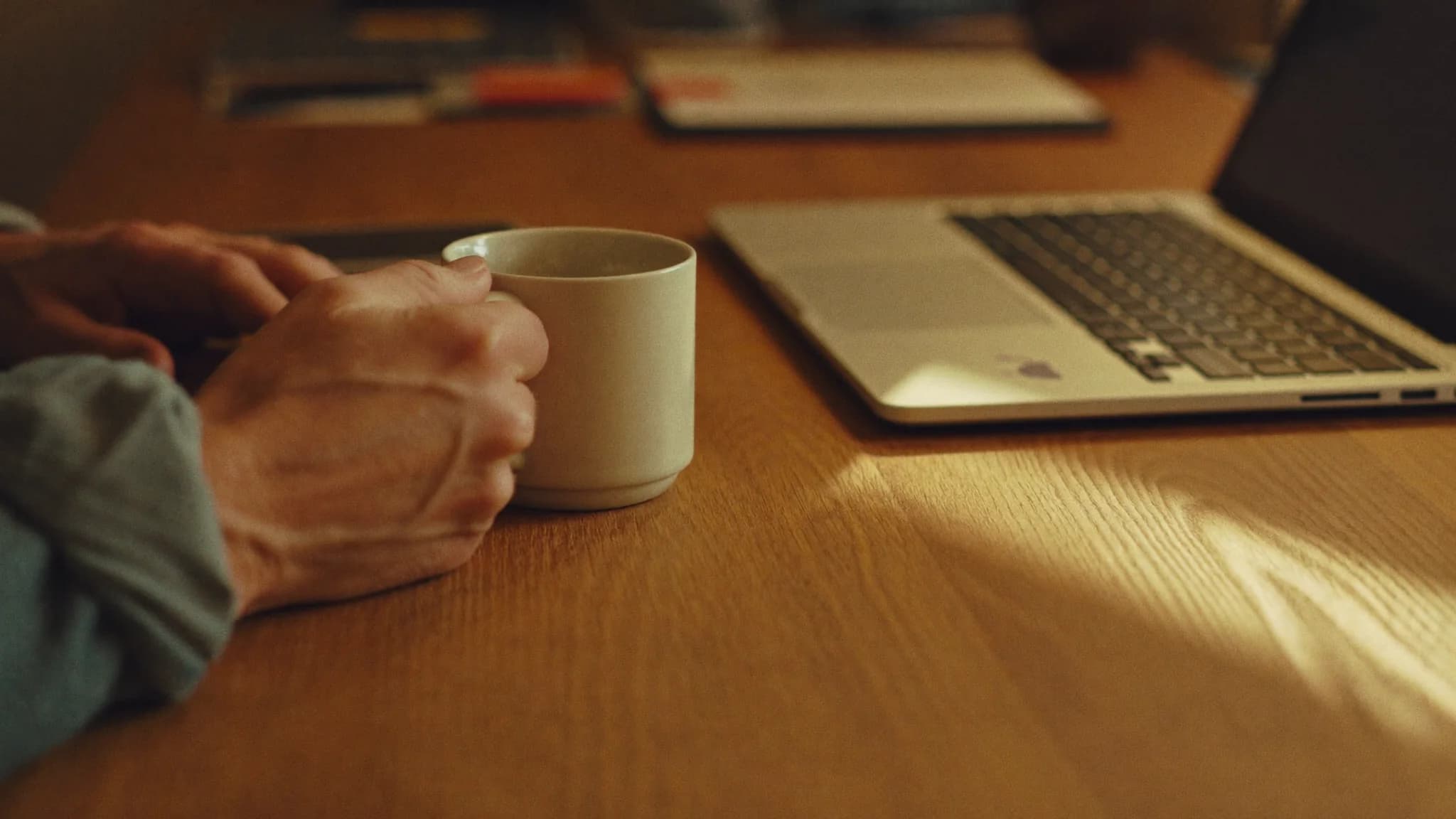 A close view of a home-services owner holding a coffee mug beside a laptop on a warm oak desk in late-afternoon light, weighing a software decision.