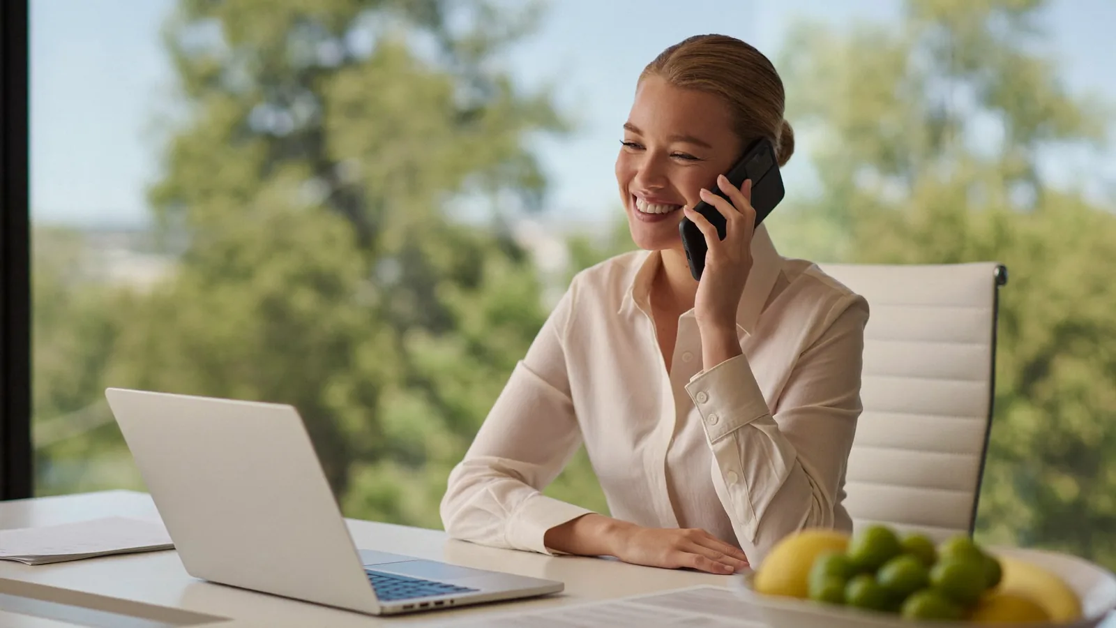 A leasing agent smiling while talking on the phone at a bright office desk in warm afternoon light.