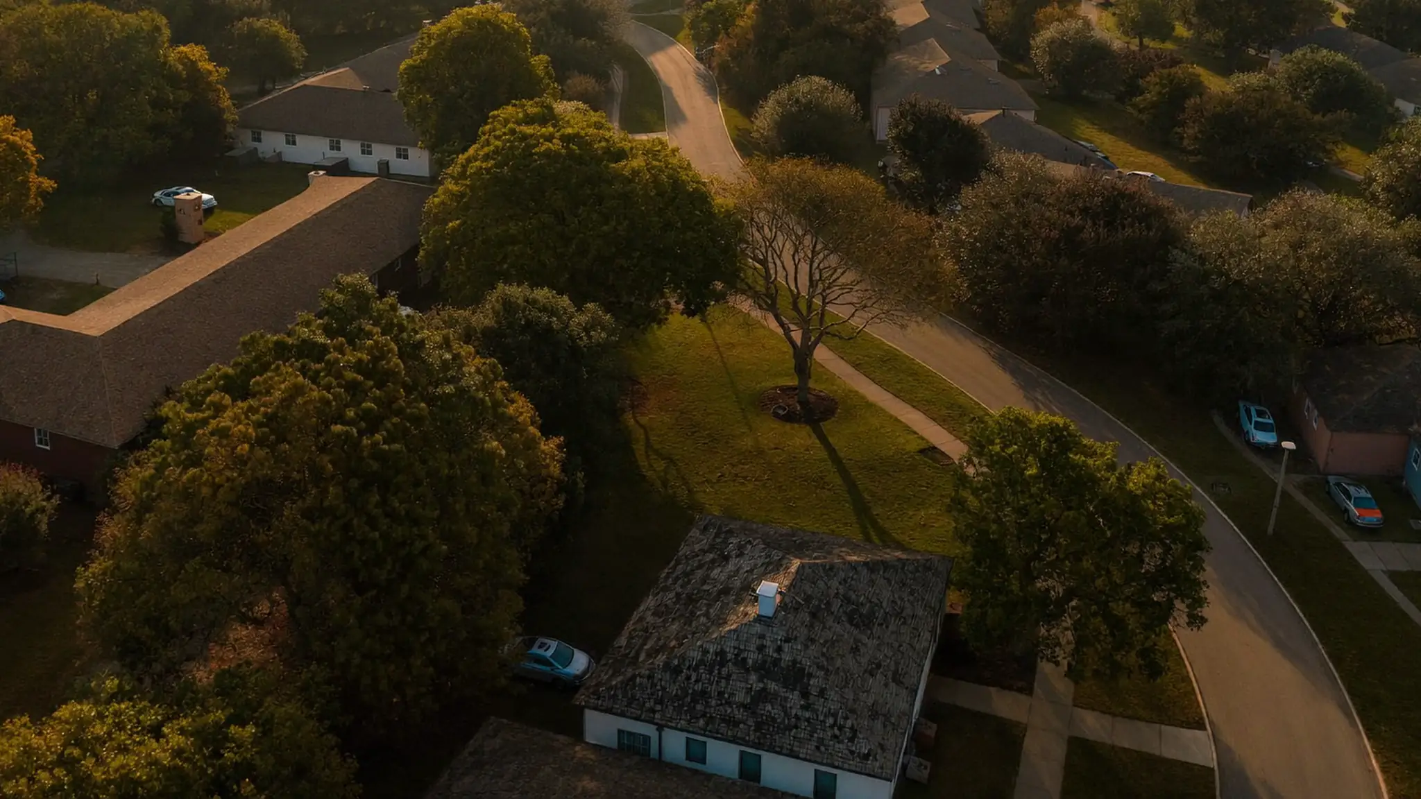 An elevated aerial view of a suburban neighborhood grid of houses, streets and lawns at golden hour.
