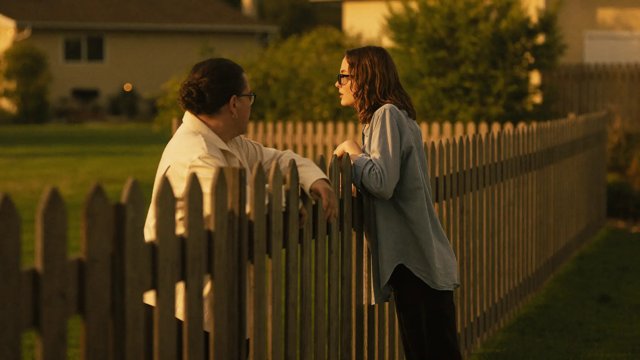 Two neighbors chatting over a low backyard fence in a suburban yard at golden hour.