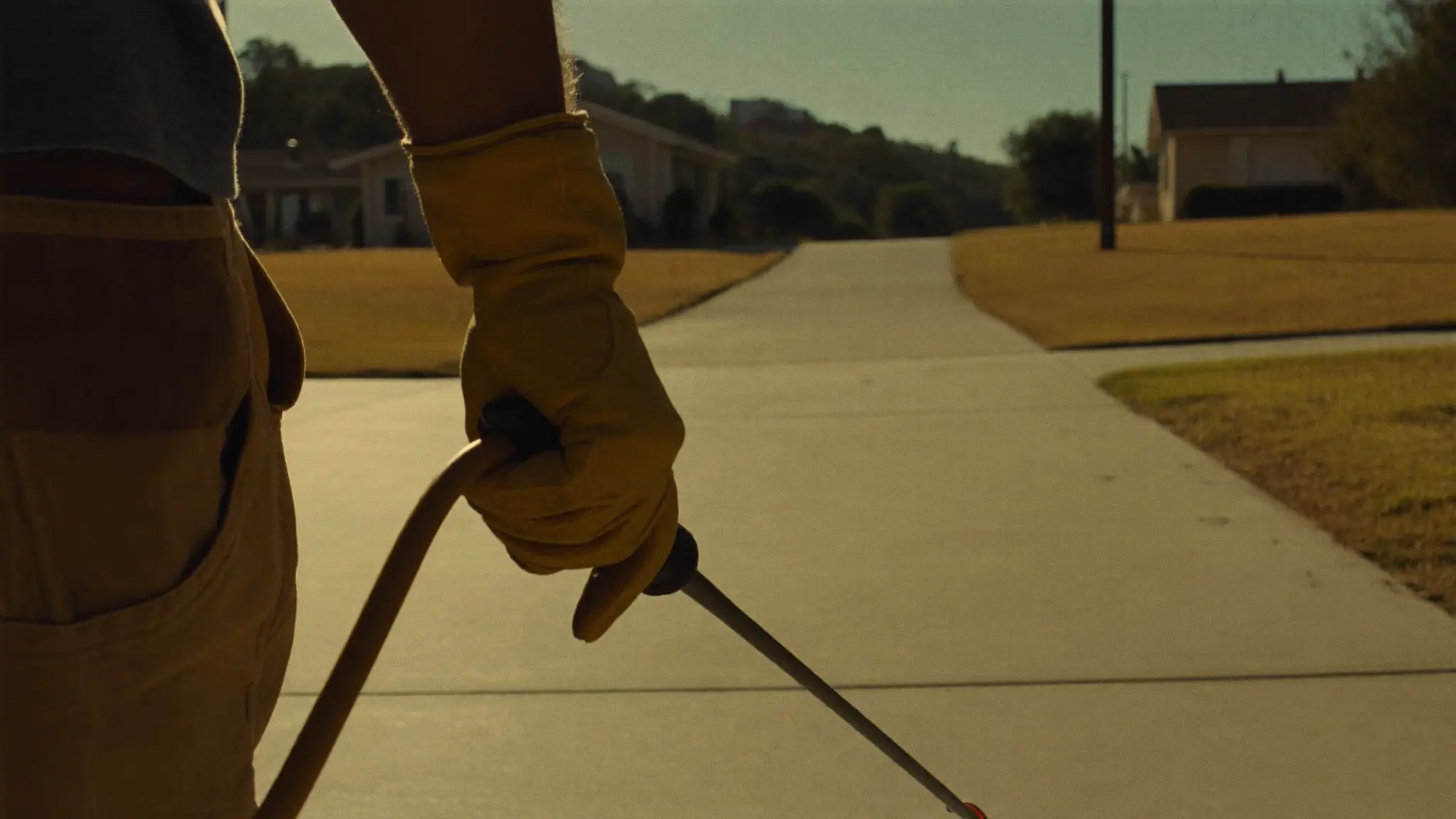 A pest control technician's gloved hand carrying a sprayer wand up a suburban driveway in warm morning light.