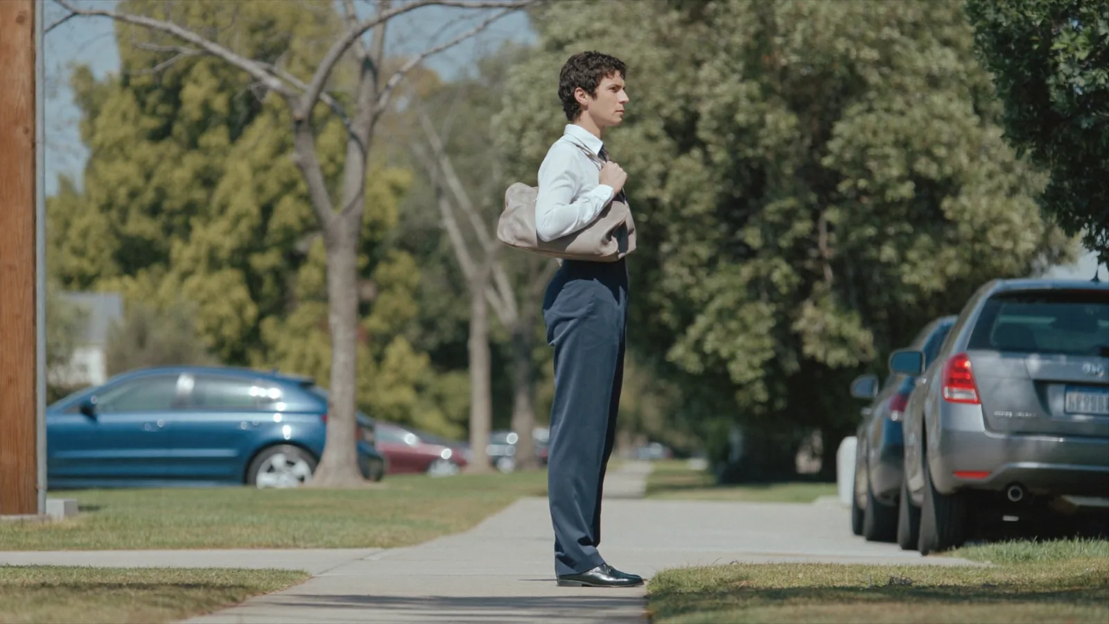 A field salesperson pausing on a quiet suburban sidewalk in daylight, composed and determined, bag on the shoulder.