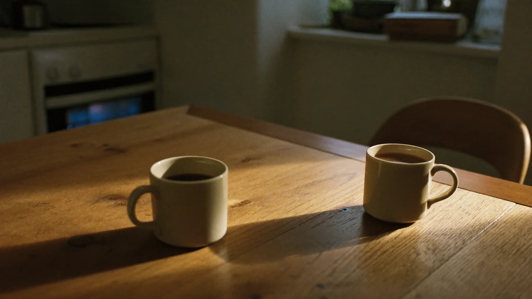 Two plain coffee mugs on a worn wooden kitchen table in warm late-afternoon light, an in-home sales conversation paused.