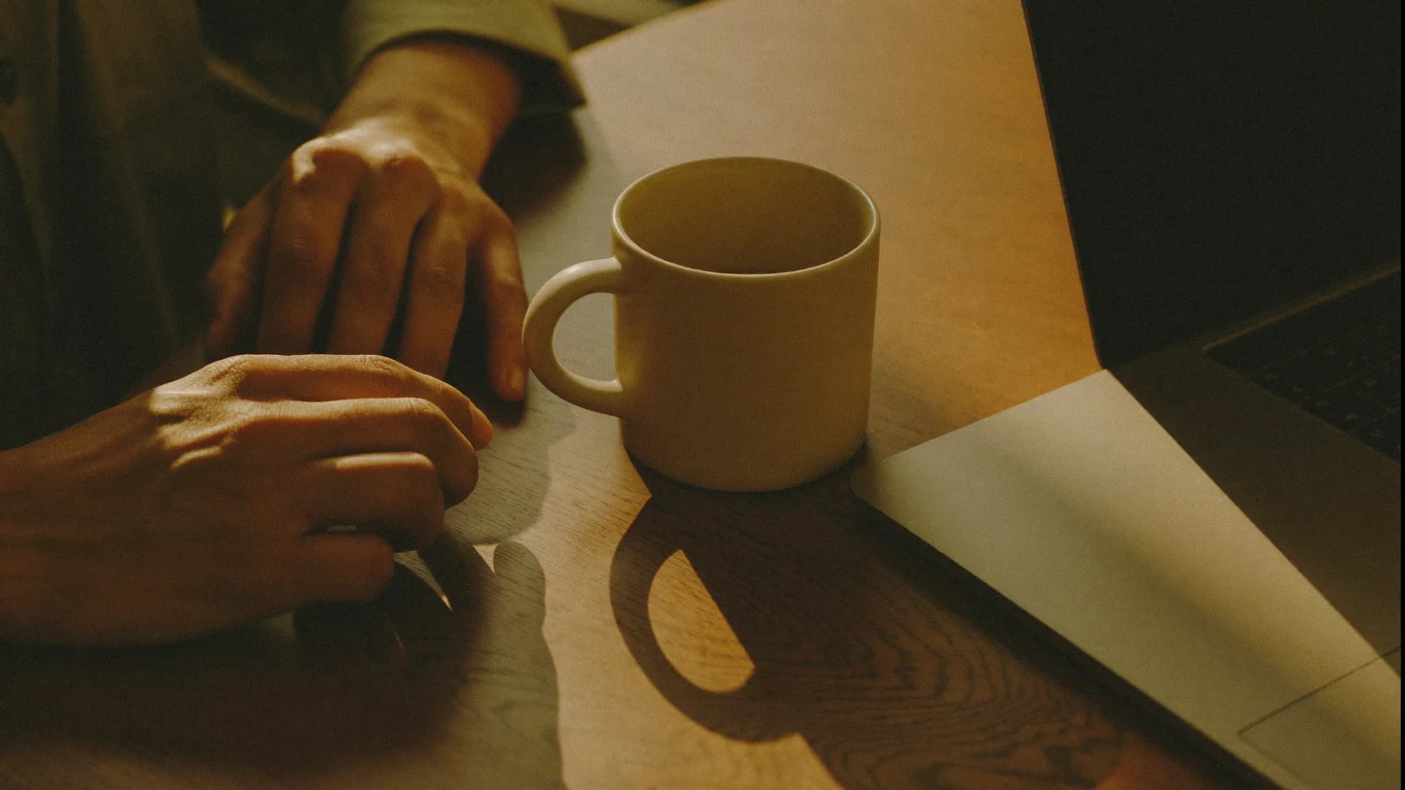 A home services business owner's hands resting on a kitchen table beside a coffee mug and a closed laptop in warm morning light.