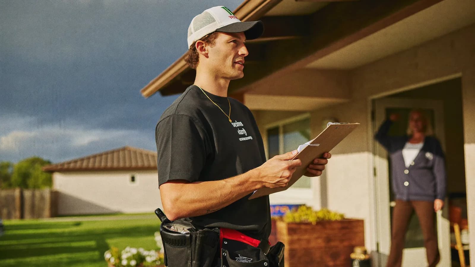 A roofing contractor with a clipboard talking to a homeowner on a front porch under a clearing storm sky.