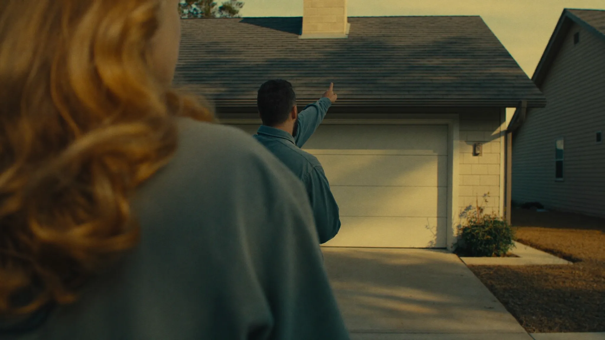 A roofing contractor and a homeowner talking in a driveway, the contractor gesturing toward the roof in afternoon daylight.