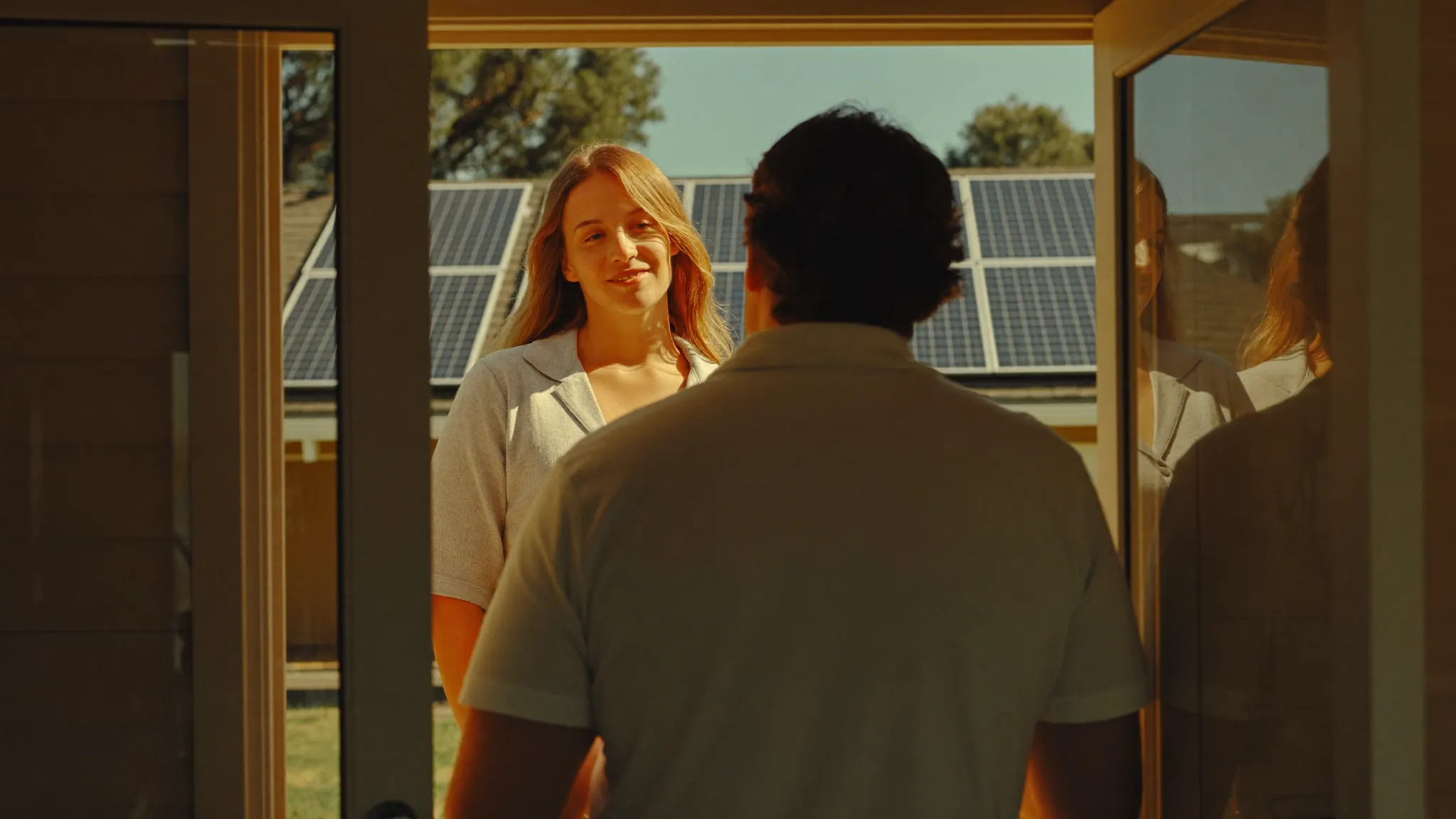 A solar sales rep in a plain polo talking with a homeowner on a sunny front porch, solar panels on a neighboring roof.