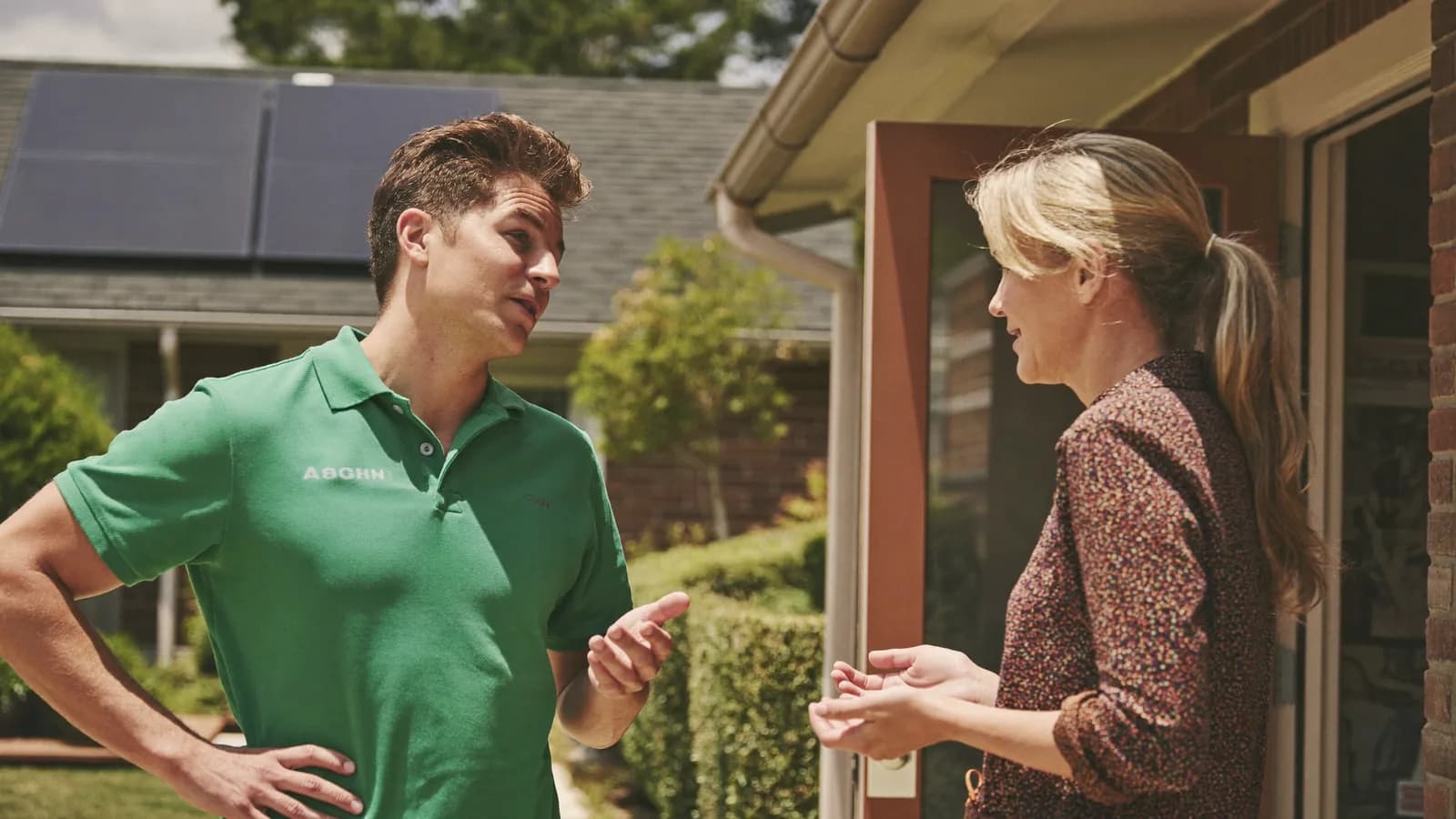 A solar sales rep in a plain polo talking with a homeowner on a sunny front porch, solar panels on a neighboring roof.