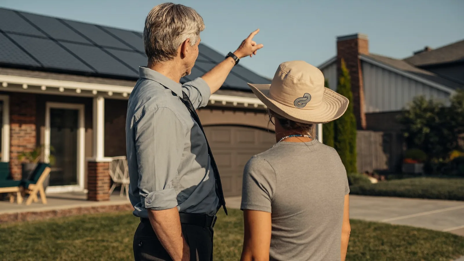 A solar sales rep and a homeowner standing in a driveway looking up toward the roofline in warm afternoon light.